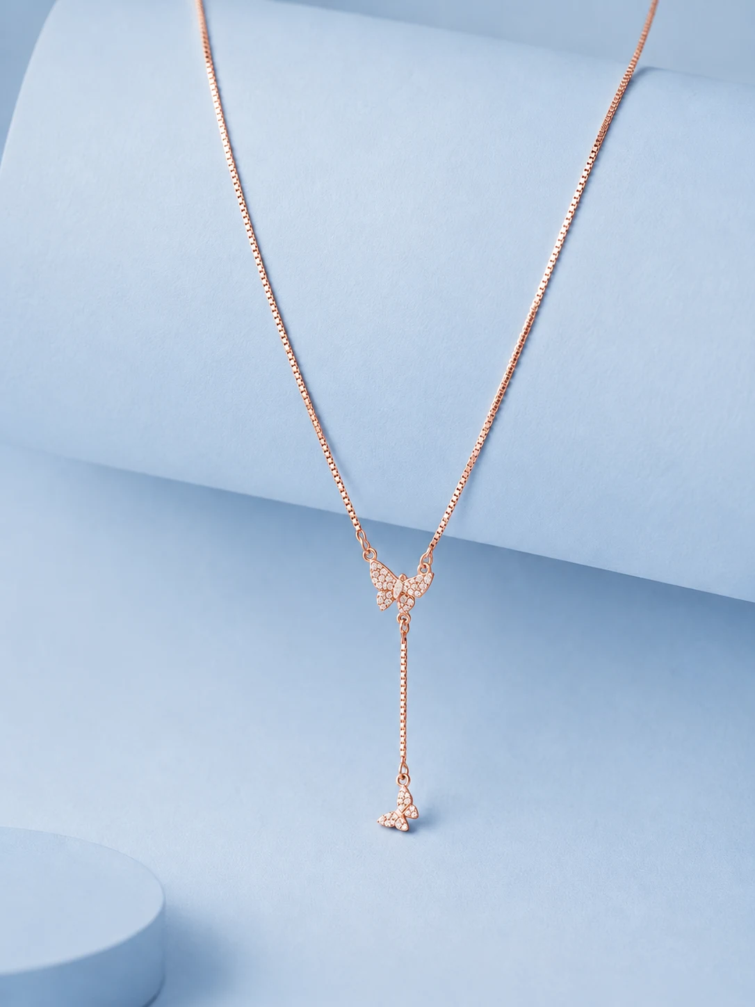 Website image rose gold butterfly Rose gold butterfly drop necklace with zircon detailing displayed on a soft pastel blue background.