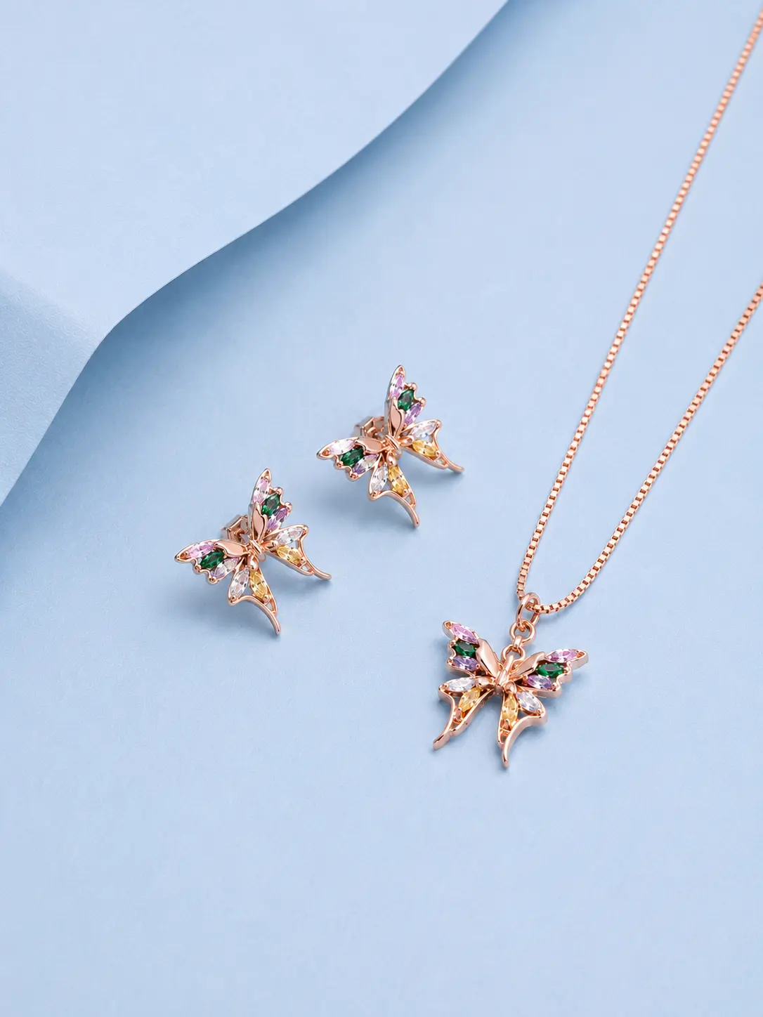 butterfly pendant earring rosegold Rose gold butterfly pendant necklace with matching butterfly earrings featuring multicolour zircon stones displayed on a pastel blue background.