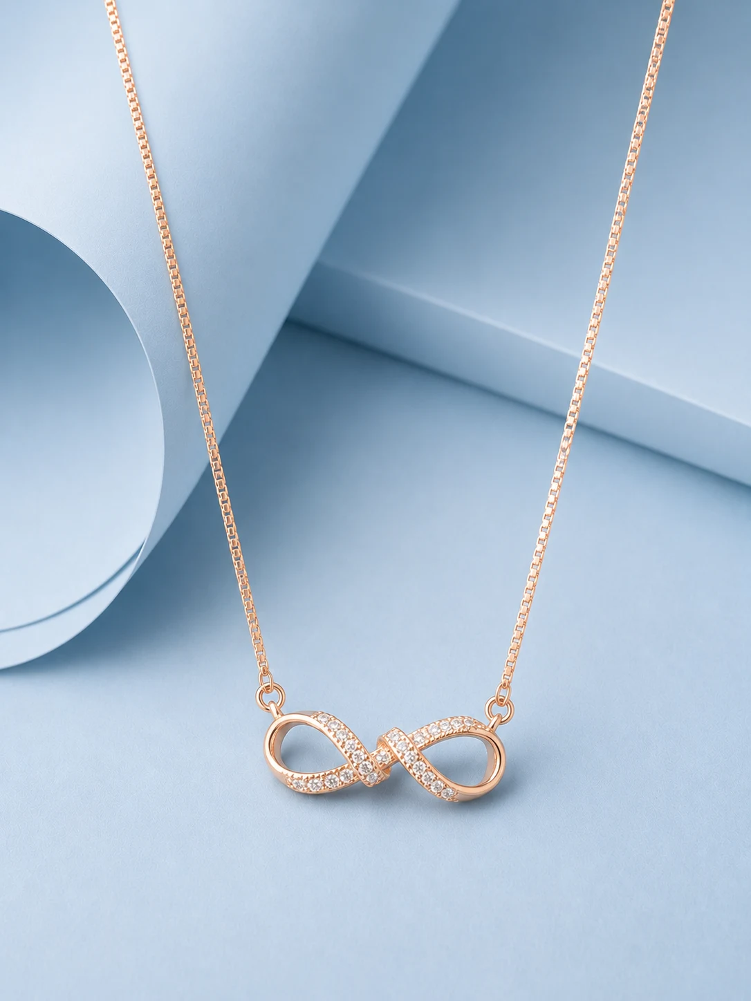 infinity @2 braid rose gold web (2) Rose gold infinity necklace with intertwined zircon-studded design displayed on a pastel blue background.