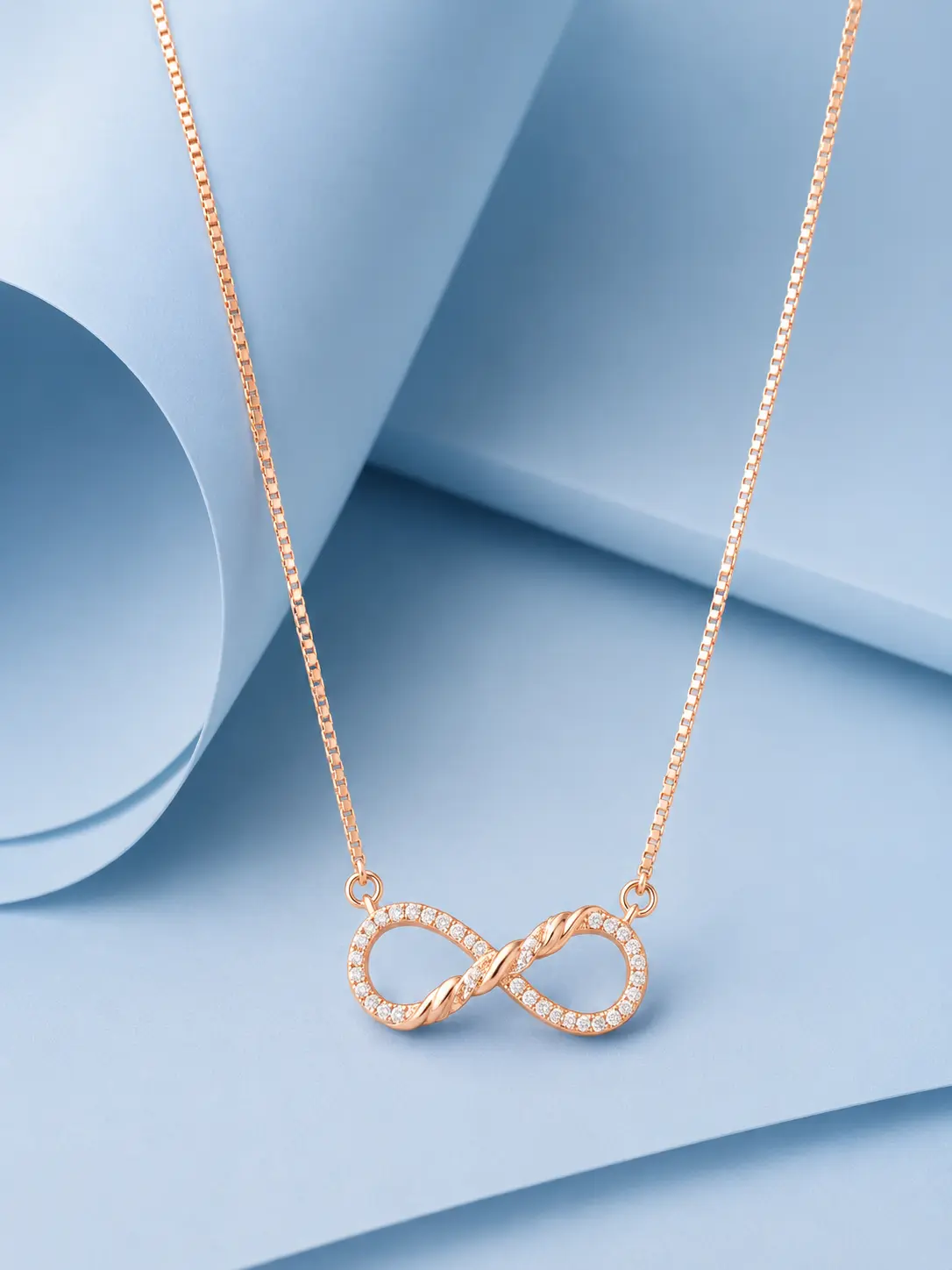 infinity braid rose gold web Rose gold twisted infinity necklace with zircon detailing displayed on a pastel blue geometric background.