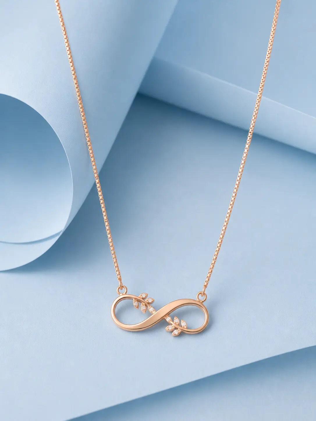 leef infinity Rose gold web Rose gold infinity necklace with delicate leaf accents and zircon detailing displayed on a pastel blue background.