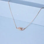 Rose gold feather-inspired necklace with zircon detailing and pearl accent displayed on a soft pastel blue background.