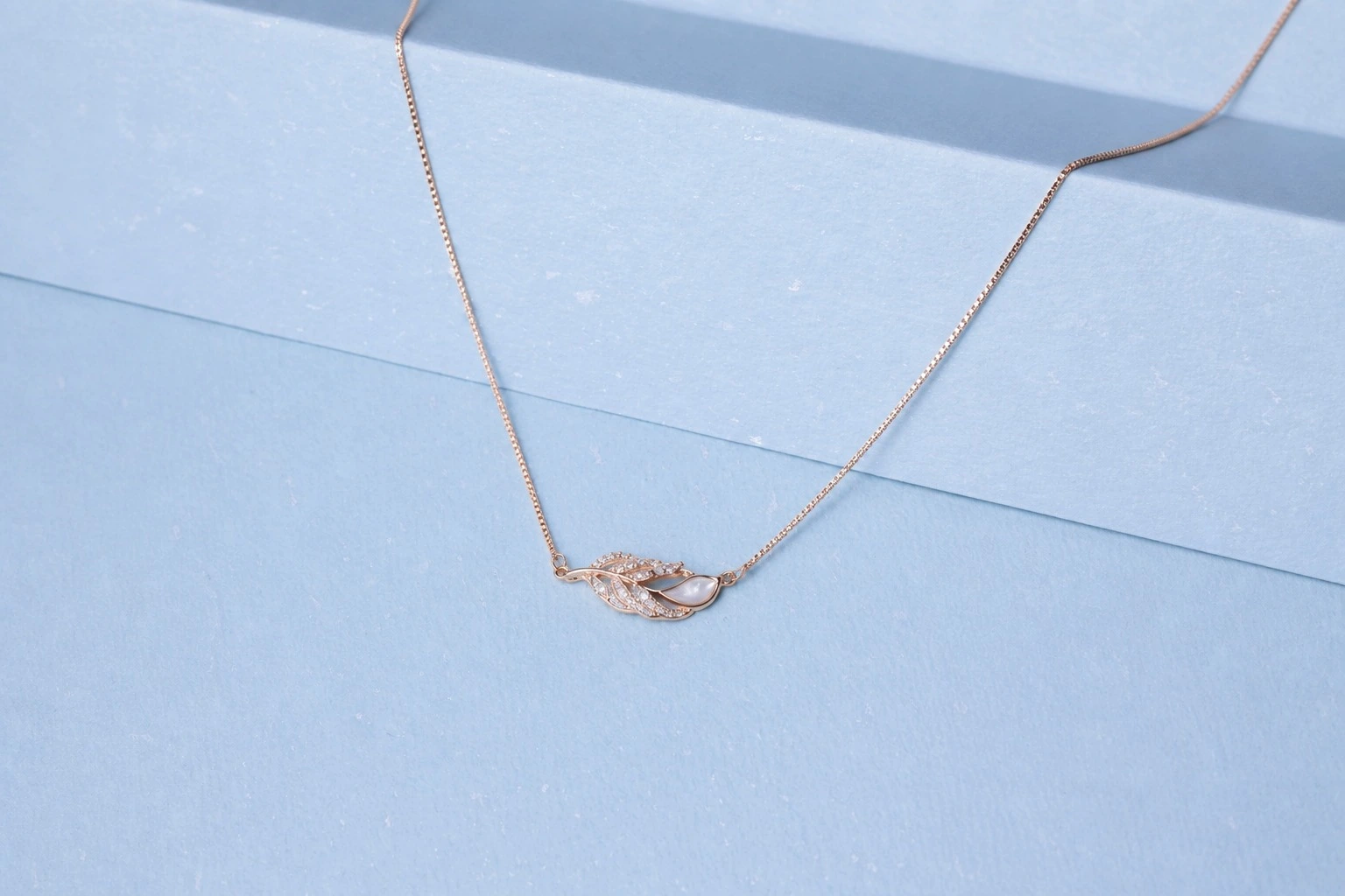 website image rose gold feather Rose gold feather-inspired necklace with zircon detailing and pearl accent displayed on a soft pastel blue background.