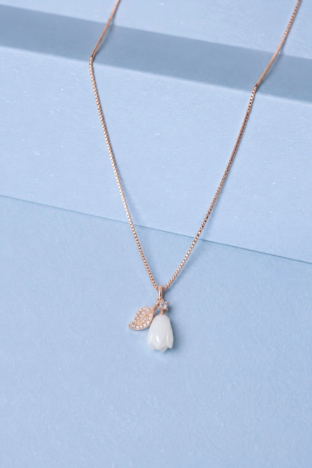 website image rosegold rose pendant Rose gold floral pendant necklace with ivory flower charm and zircon detailing displayed on a soft pastel blue background.