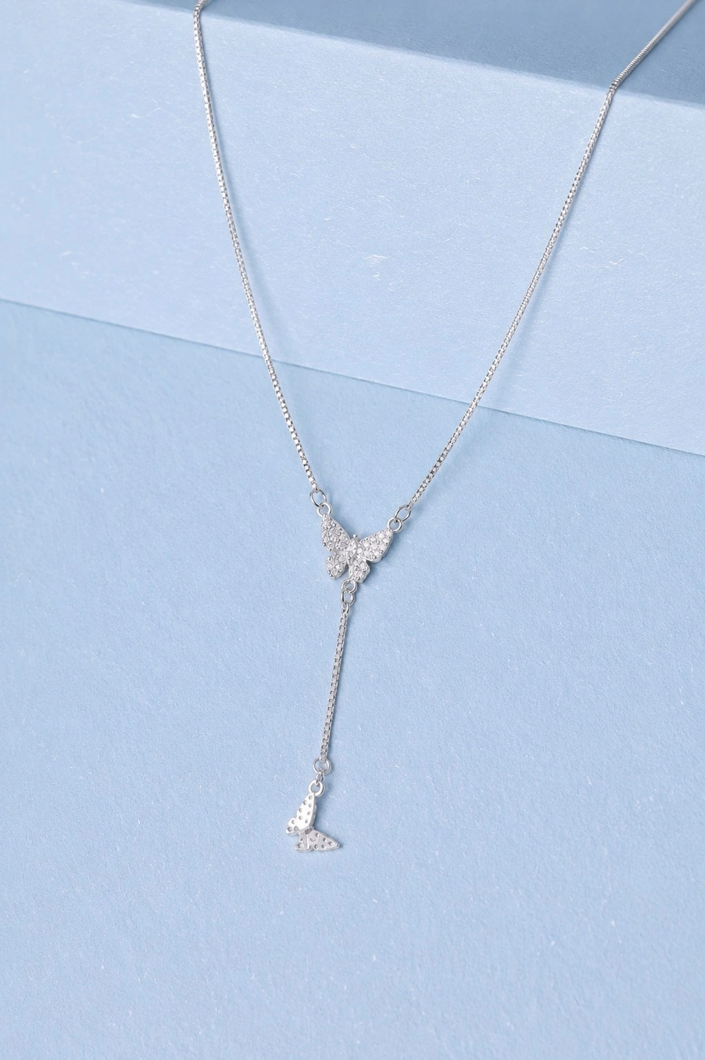 wesite imagesilver butterfly Sterling silver butterfly drop necklace with zircon detailing displayed on a soft pastel blue background.