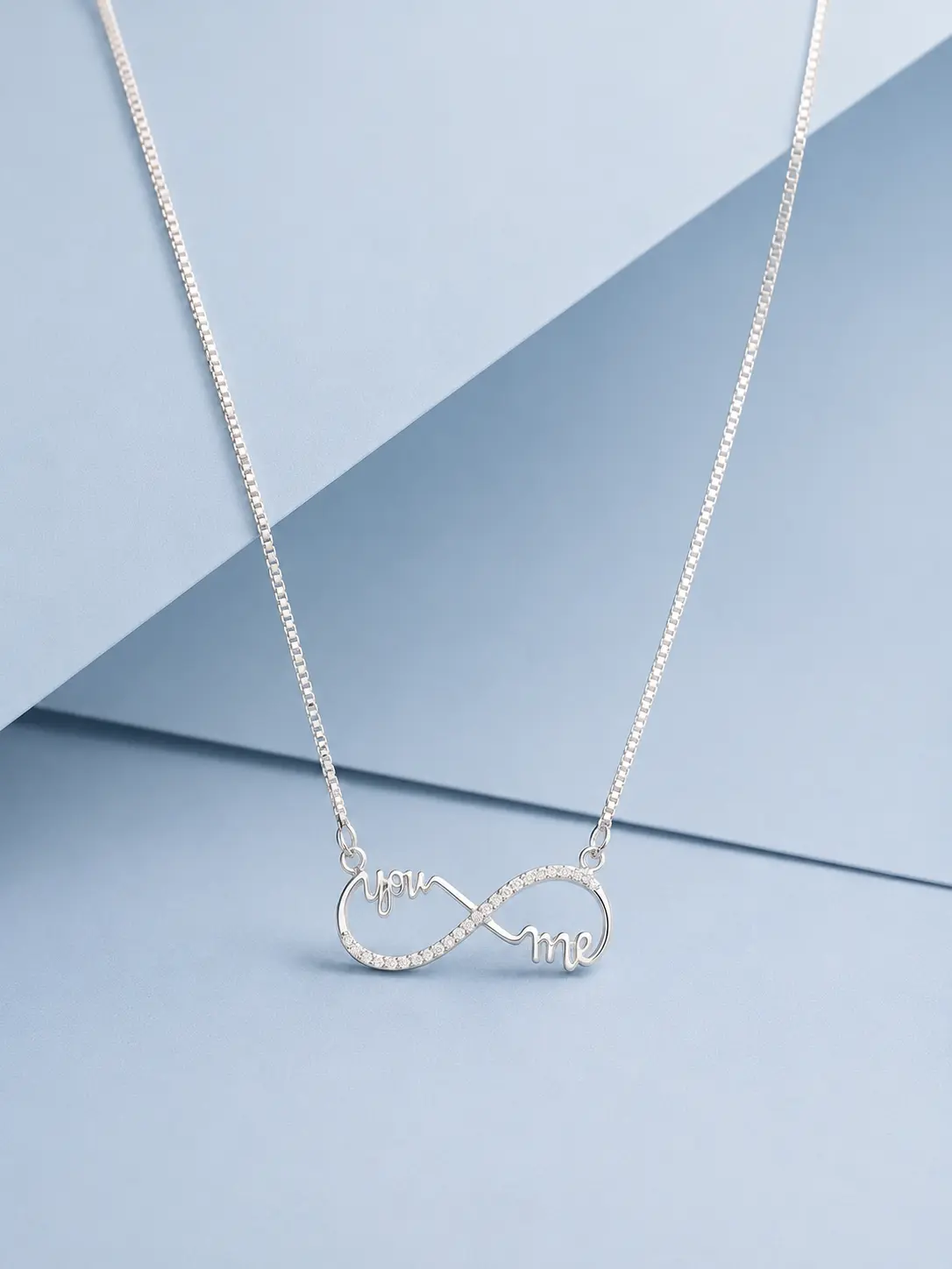 you & me silver web Sterling silver infinity necklace with “You & Me” lettering and zircon detailing displayed on a pastel blue luxury background.
